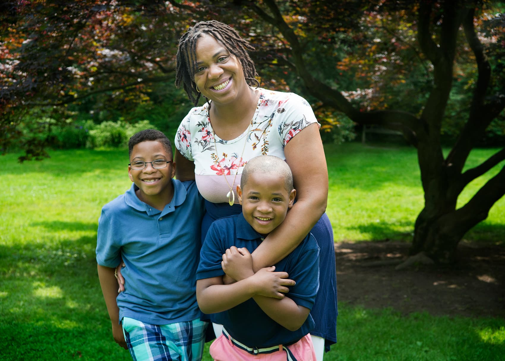 Smiling woman hugs two young boys while standing outside in a sunny park