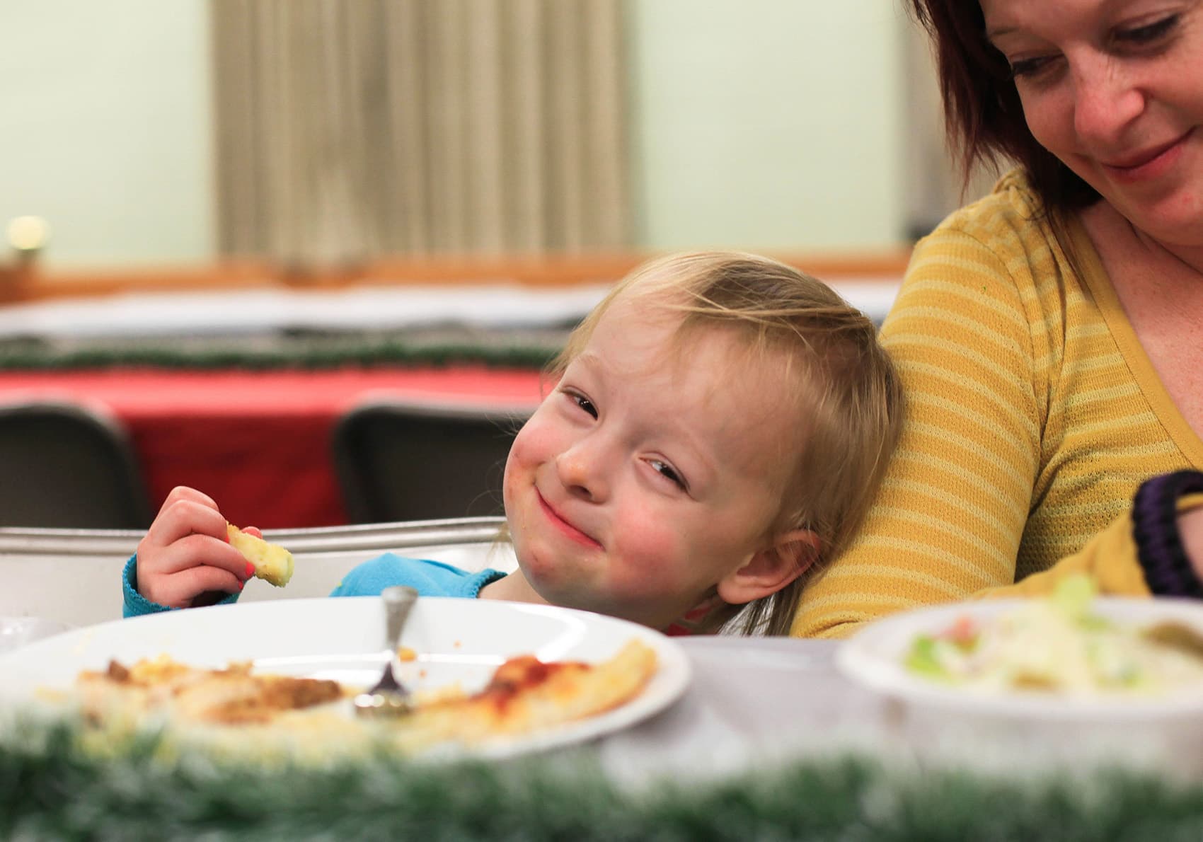 Smiling child leans on woman’s arm while eating at a table with plates of food