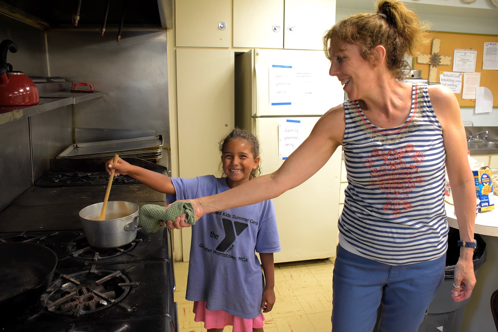 Smiling woman helps young girl stir a pot on the stove in a kitchen